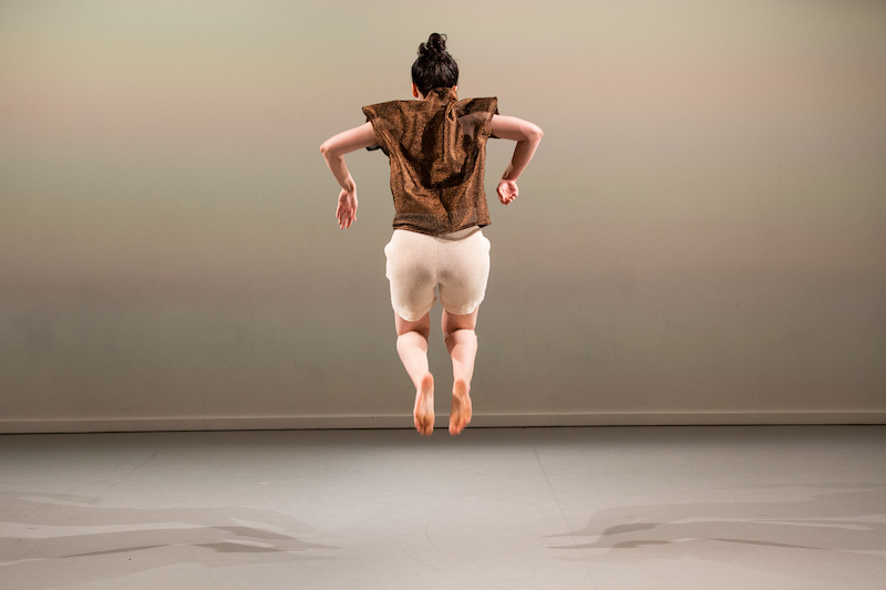 Lily Bo Shapiro jumps in the air with her feet and legs tucked underneath her. Her elbows make an almost ninety degree angle. Her back is towards the audience